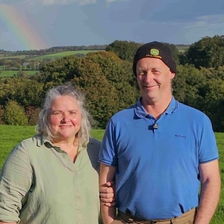 Ben and Denise smiling together outdoors on their farm, with a rainbow visible behind them