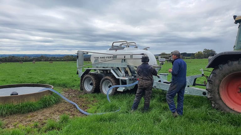 Two operators filling a Tow and Fert MultiCrop 4000 sprayer with water from a trough using a suction hose in a green paddock.