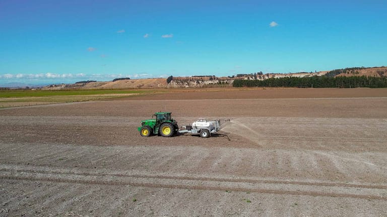 A green tractor towing a Tow and Fert Multi 2800 sprayer across a dry, open field under a blue sky.