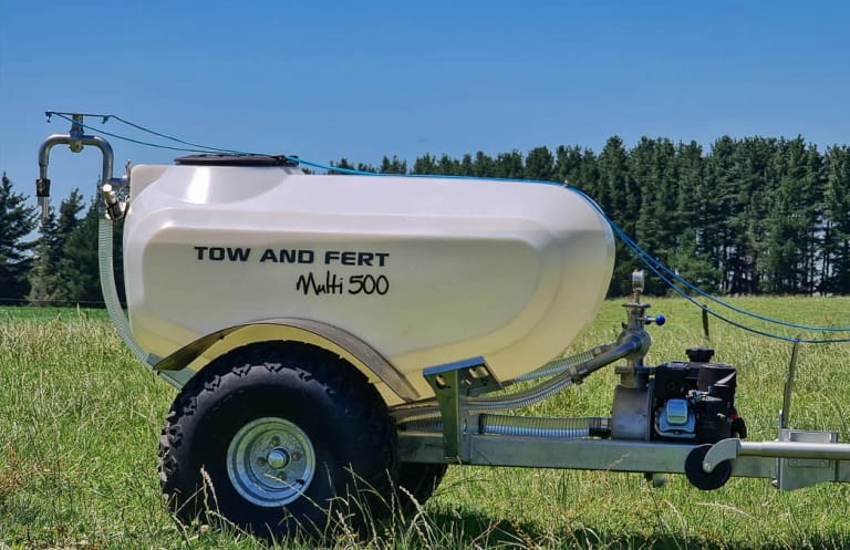 Close-up side view of a Tow and Fert Multi 500 tow-behind sprayer in a green pasture, showing its white tank, pump, hoses and large all-terrain wheel.