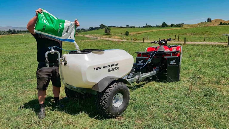 A farmer pouring granular fertiliser into the tank of a Tow and Fert Multi 500 sprayer attached to a quad bike in a green pasture.
