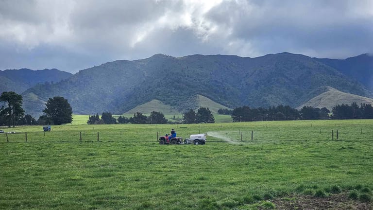 A farmer riding a red quad bike towing a Tow and Fert sprayer across a green pasture, spraying liquid fertiliser with mountains and a cloudy sky in the background.