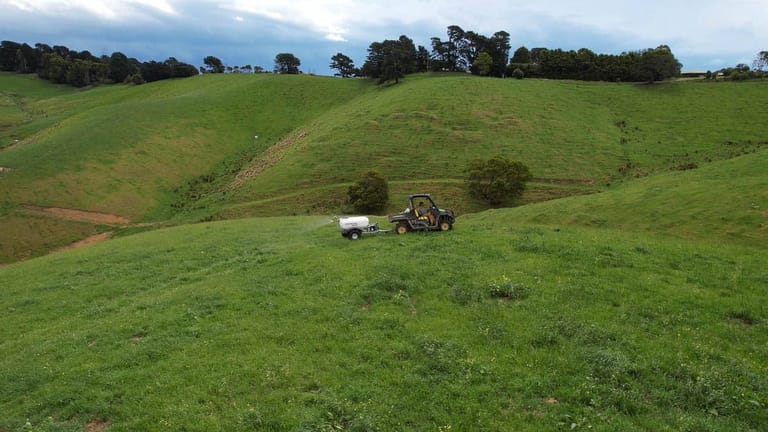 A farmer driving a utility vehicle towing a Tow and Fert sprayer across a green, hilly pasture, applying liquid fertiliser with rolling hills in the background.