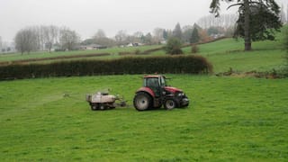 A red tractor towing a Tow and Fert Multi 1000 sprayer across a green pasture, with rolling hills and trees in the background.