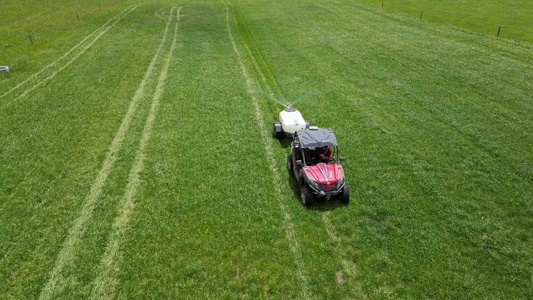 Aerial view of a red utility vehicle towing a Tow and Fert sprayer across a green pasture, applying liquid fertiliser to the grass.