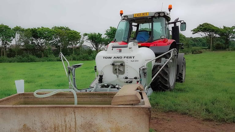 Rear view of a Tow and Fert Multi 1200 sprayer attached to a red tractor, drawing water through a suction hose to fill its tank.