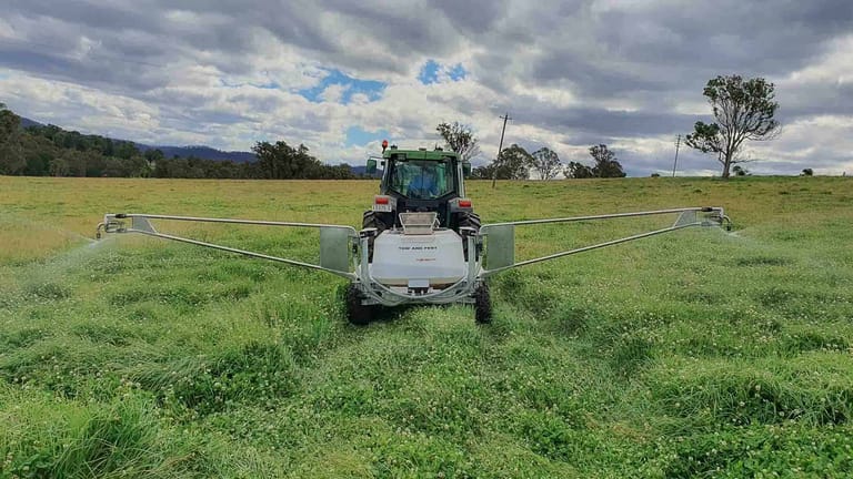 Rear view of a tractor towing a Tow and Fert Multi 1000 sprayer across a green pasture, applying liquid fertiliser through spray booms on both sides.