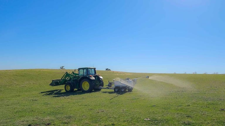 A green tractor towing a Tow and Fert sprayer across a grassy hillside, applying liquid fertiliser through an spray boom under a clear blue sky.