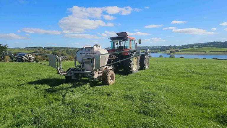 A tractor towing a Tow and Fert Multi 1200 sprayer across a green pasture under a blue sky.
