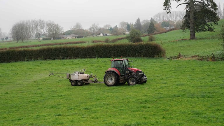 A red tractor towing a Tow and Fert Multi 1000 sprayer across a green pasture, with rolling hills and trees in the background.