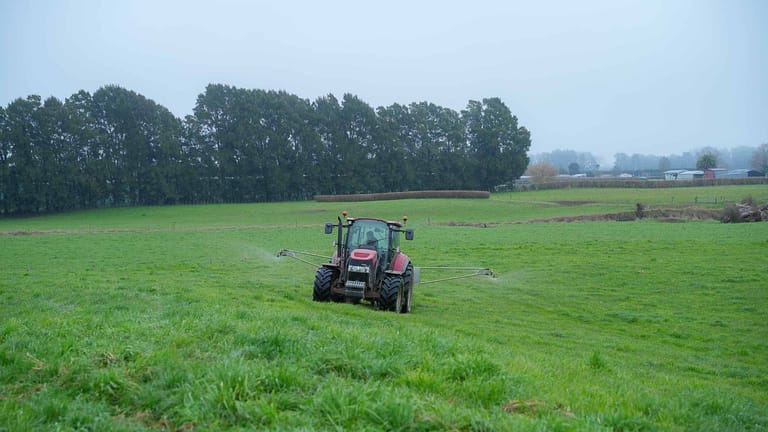 A red tractor towing a Tow and Fert Multi 1000 sprayer across a green pasture, applying liquid fertiliser through extended spray booms on both sides.