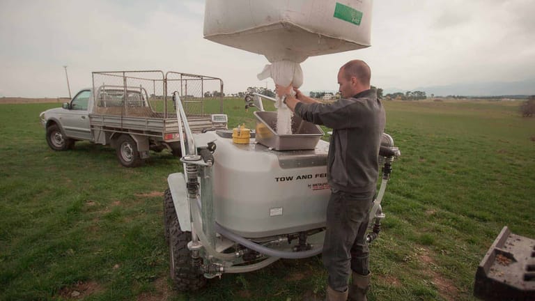 A farmer pouring powdered fertiliser from a bulk bag into the hopper of a Tow and Fert Multi 1000 sprayer in a green pasture, with a utility truck in the background.