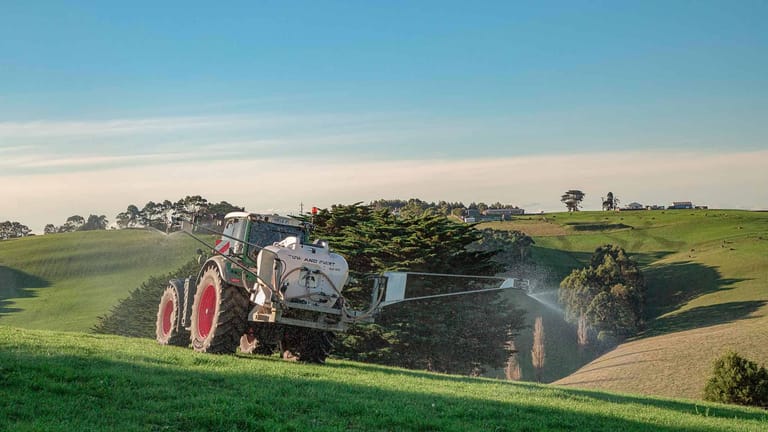 A red tractor towing a Tow and Fert Multi 1200 sprayer along a rural farm track, with green fields in the background.