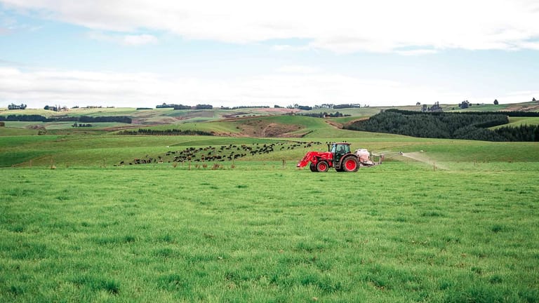A red tractor towing a Tow and Fert Multi 1200 sprayer across a green pasture on rolling farmland.